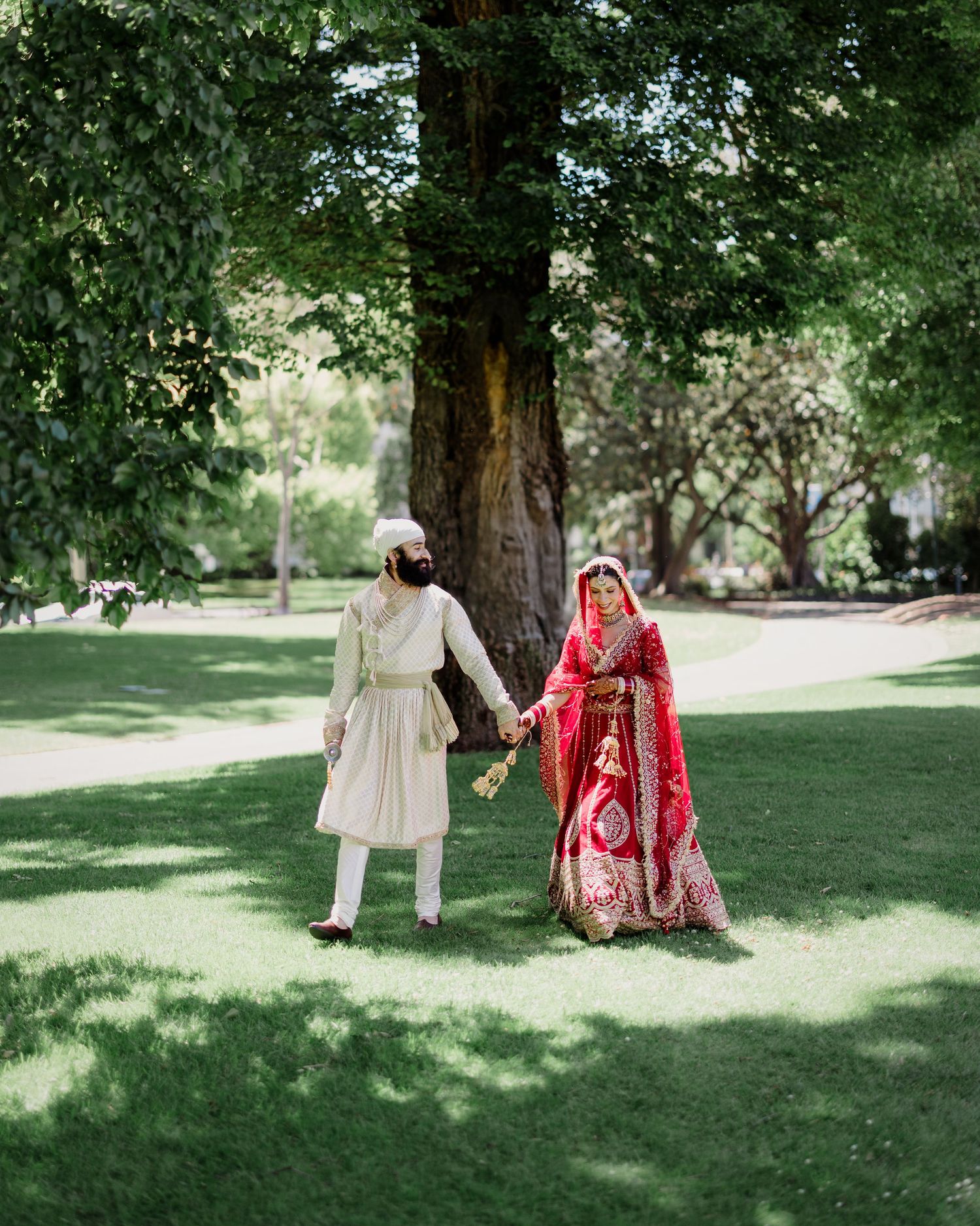 A couple in traditional wedding attire holding hands under a large tree in a lush green park setting.