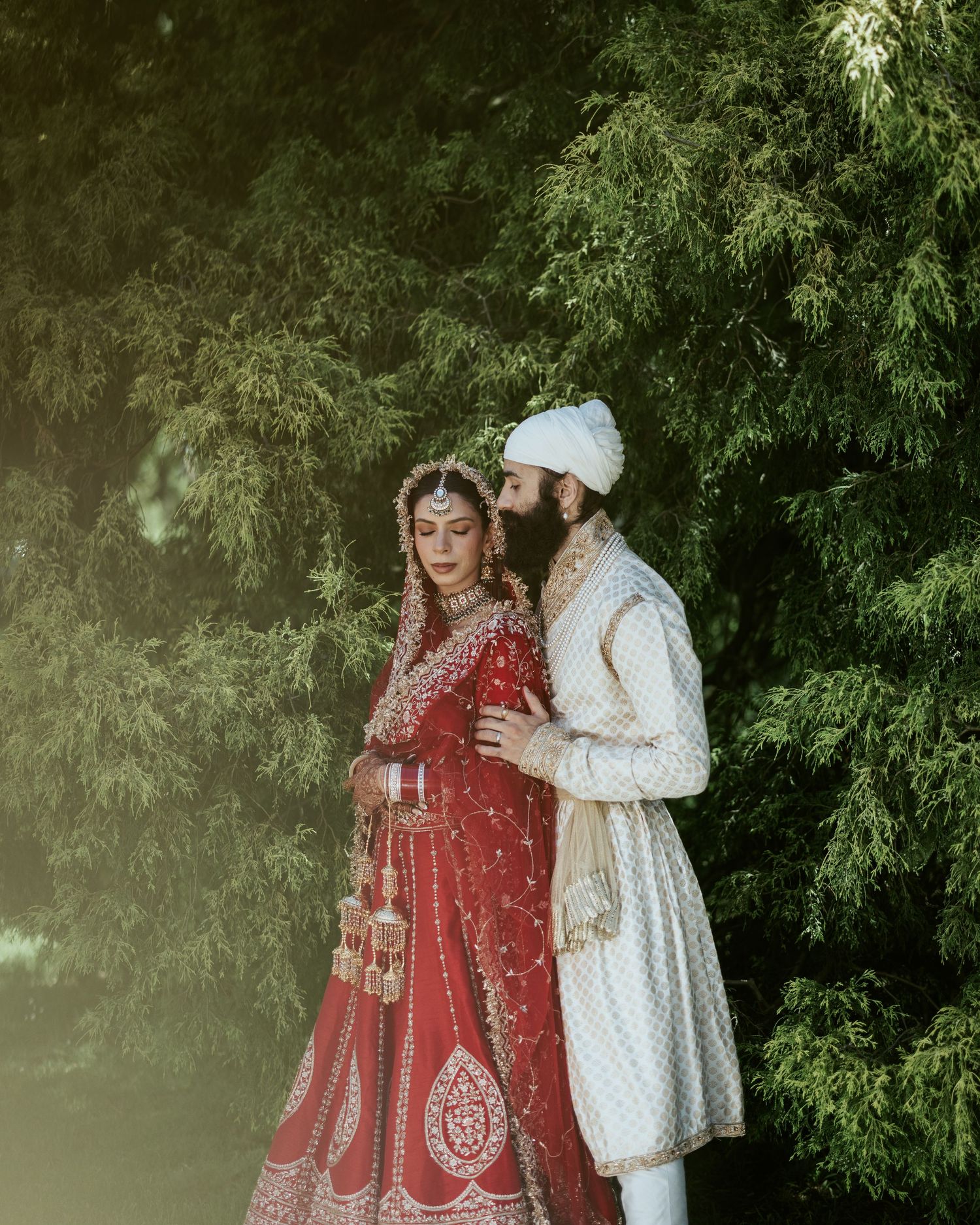A couple in traditional South Asian wedding attire poses together in a garden setting with lush green foliage background.
