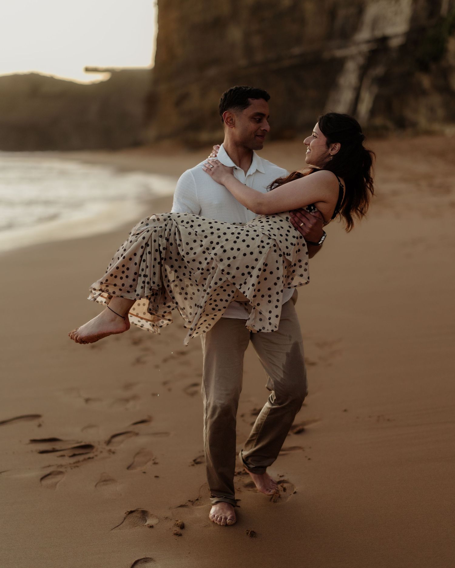 A couple shares a romantic moment on a sandy beach at sunset, as a man lifts a woman in a polka dot dress.