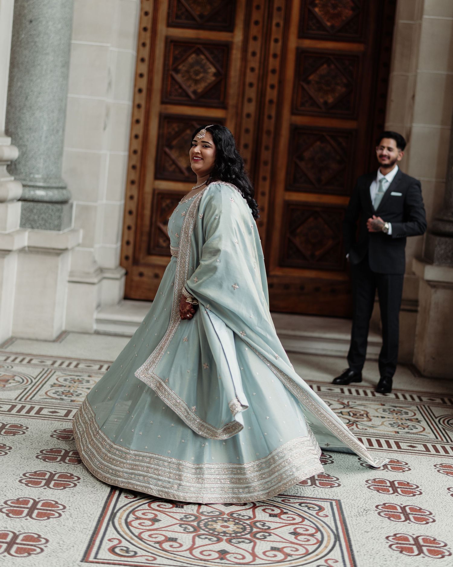 A woman twirls in a light blue embroidered lehenga outside an ornate building as a suited man watches.