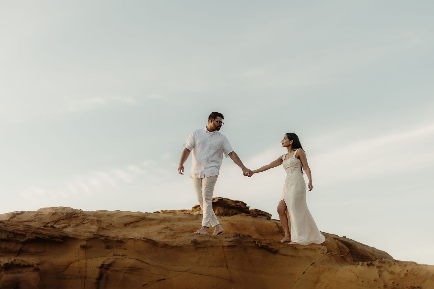 Couple in white holding hands on rocky terrain under a soft sky, romantic outdoor engagement photo.