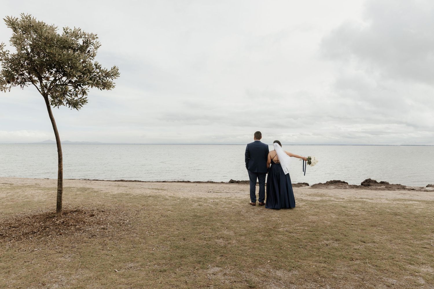 A couple sits together on a scenic beach with a lone tree in the background.