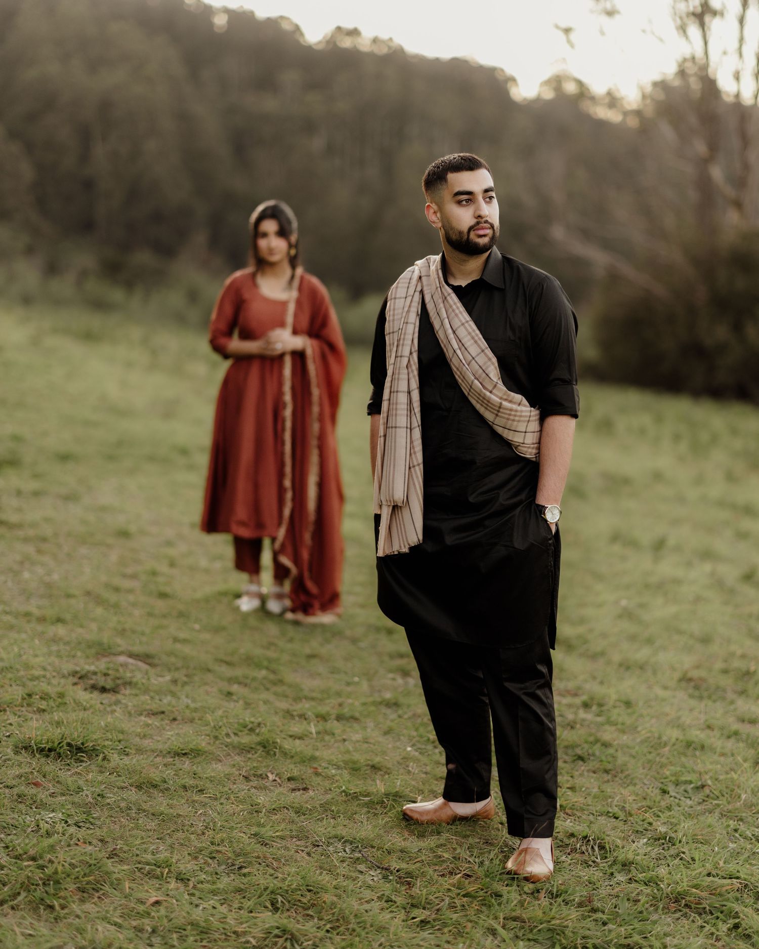 Man in black kurta with shawl stands in green field, woman in red salwar kameez blurred in background.