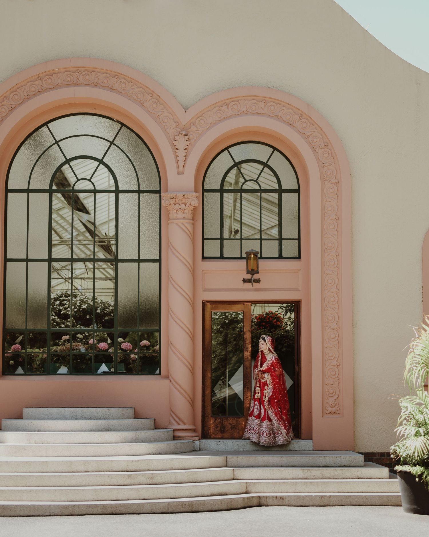 Elegant figure in a red traditional dress stands in front of pink arched doorway with ornate windows at a grand entrance.