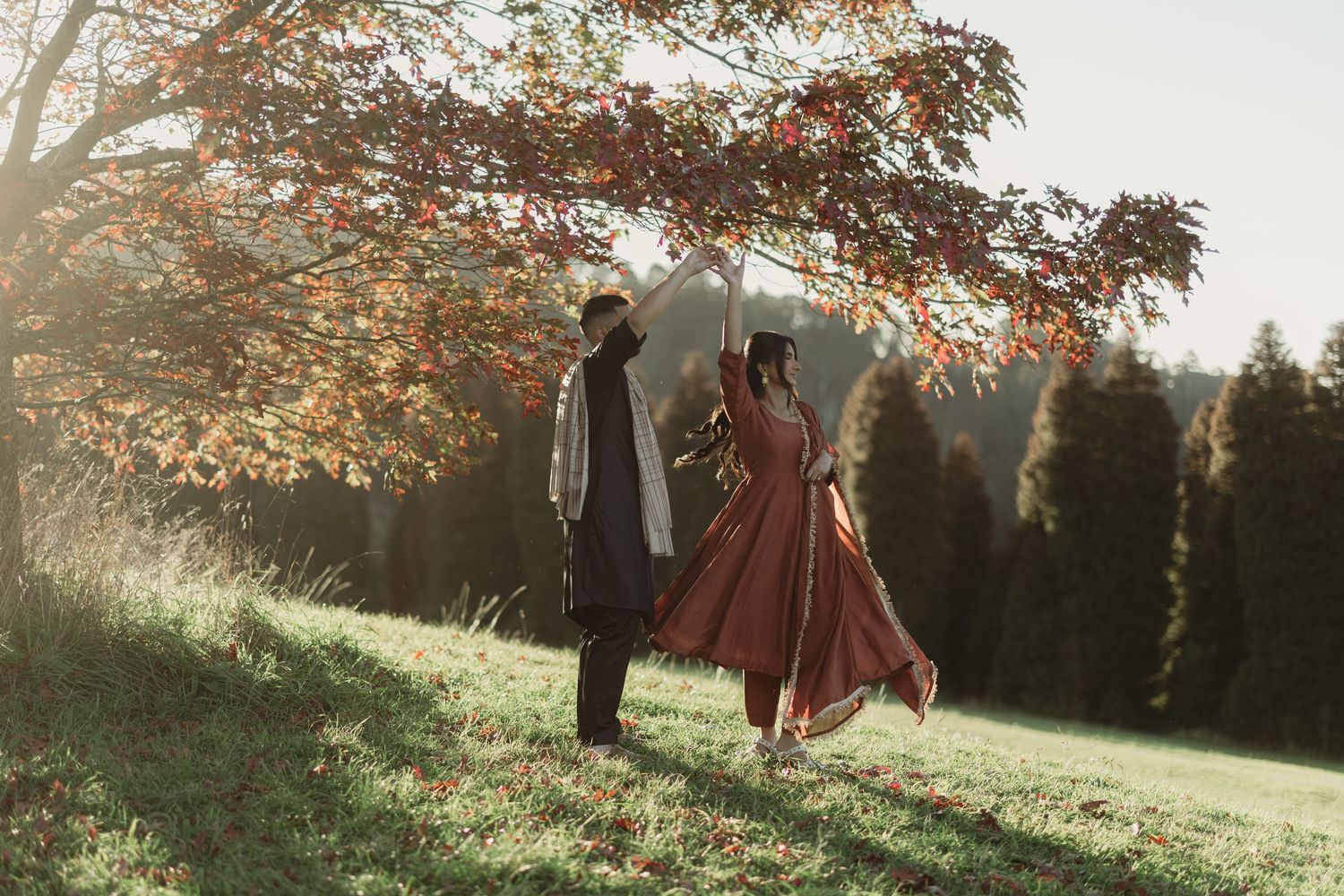 Couple in autumn forest, woman in flowing red dress, golden sunlight filtering through colorful fall foliage.