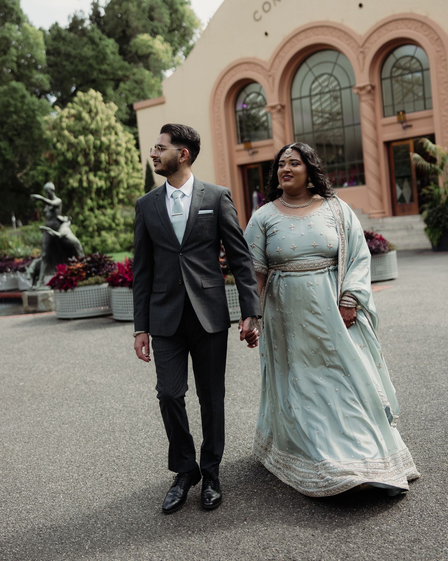 A couple in formal attire walks hand-in-hand outside an elegant venue, woman in light blue lehenga, man in dark suit.