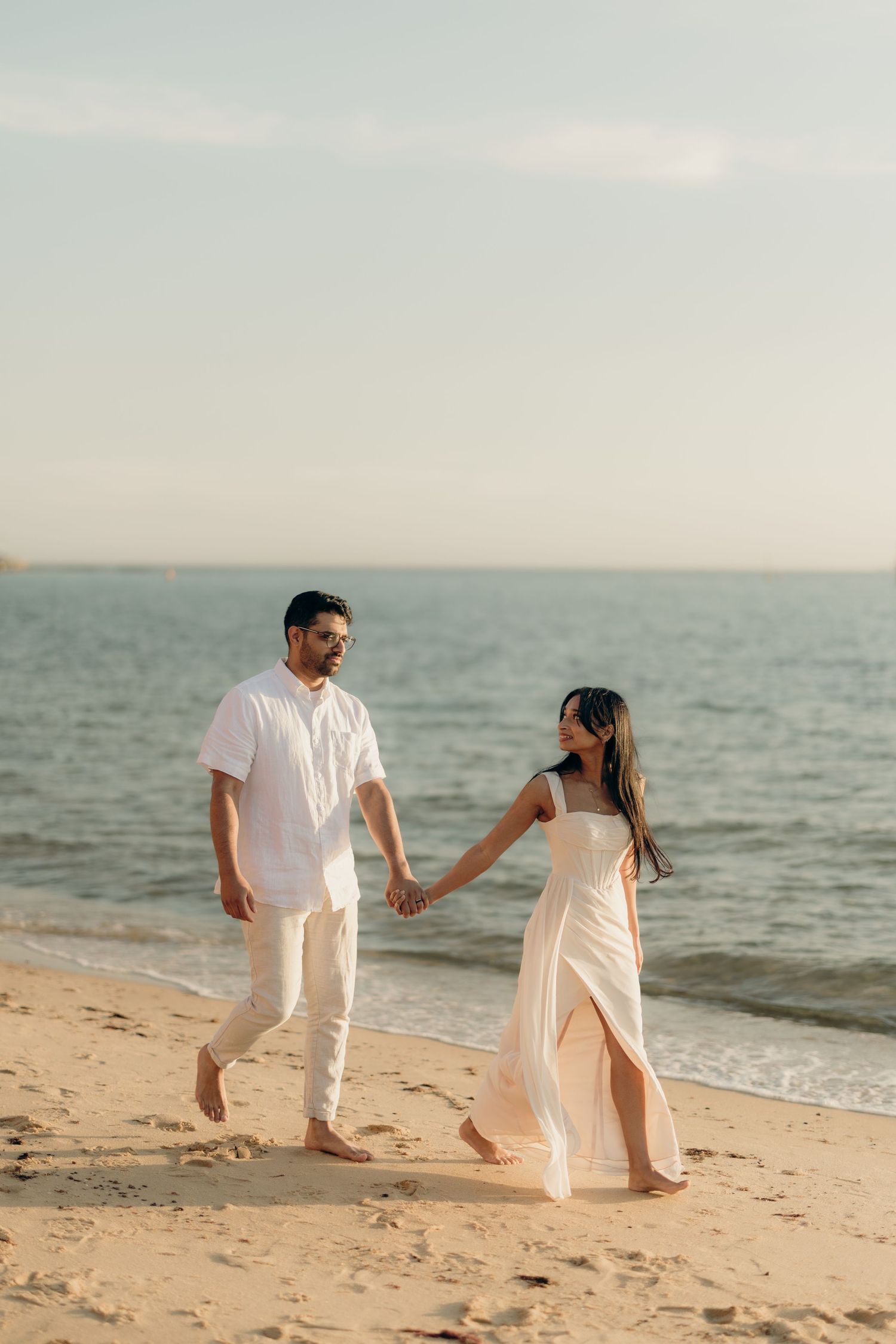 Couple in white outfits holding hands walking barefoot on a sandy beach with ocean waves in the background.