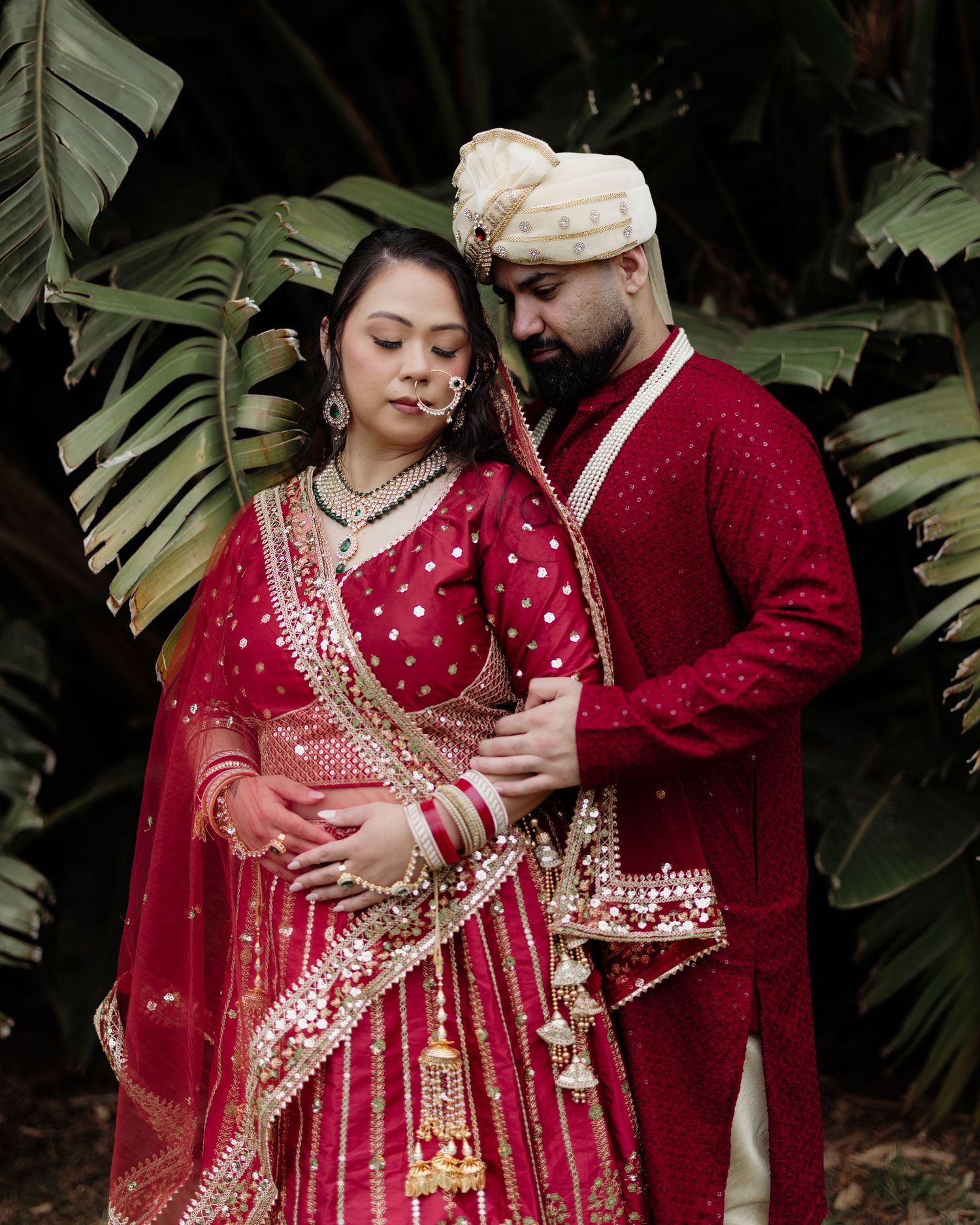 Indian bride and groom in matching red traditional wedding attire pose romantically outdoors among tropical greenery.