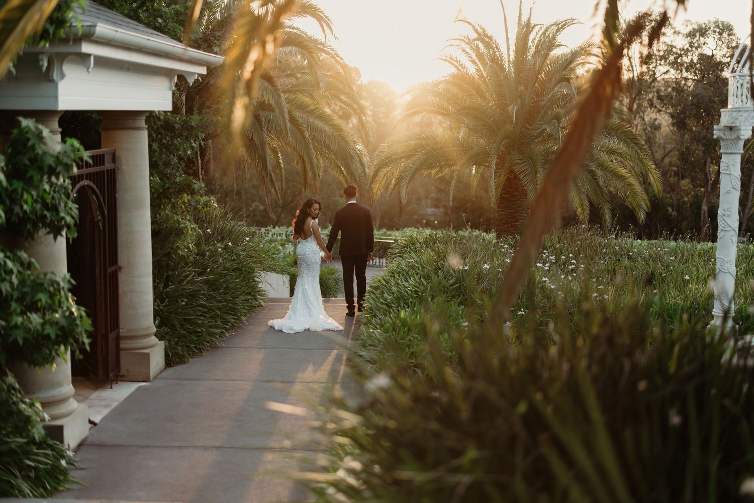 Newlyweds walking along palm-lined pathway during golden hour sunset.