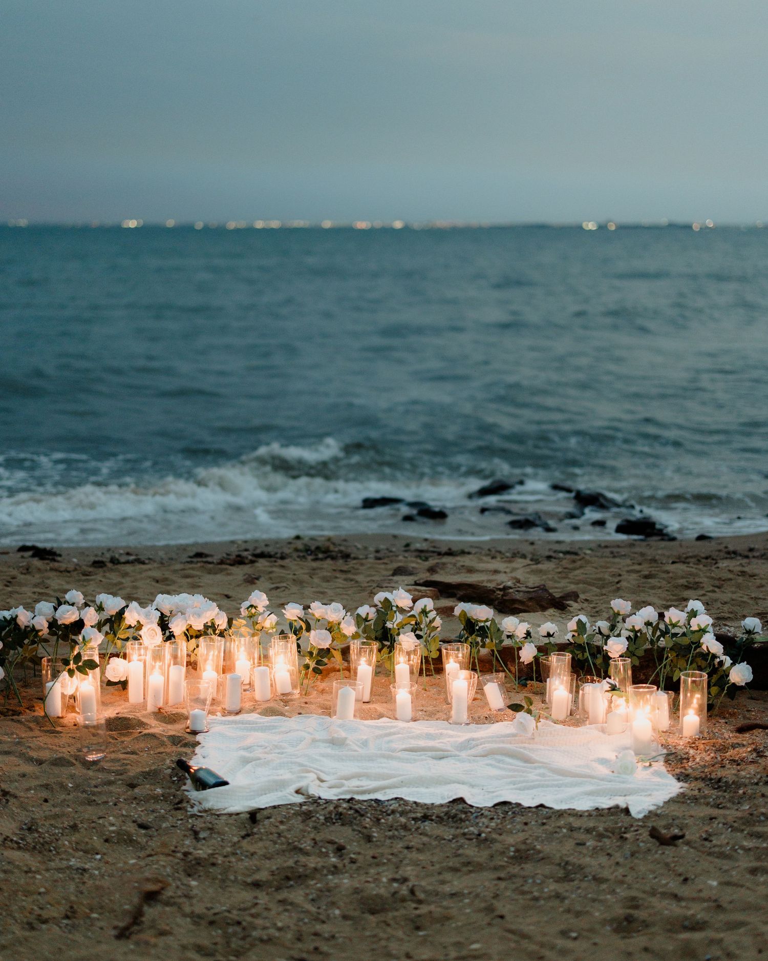 Glowing candles and white flowers create a romantic beach scene at dusk with turquoise ocean waves in the background.