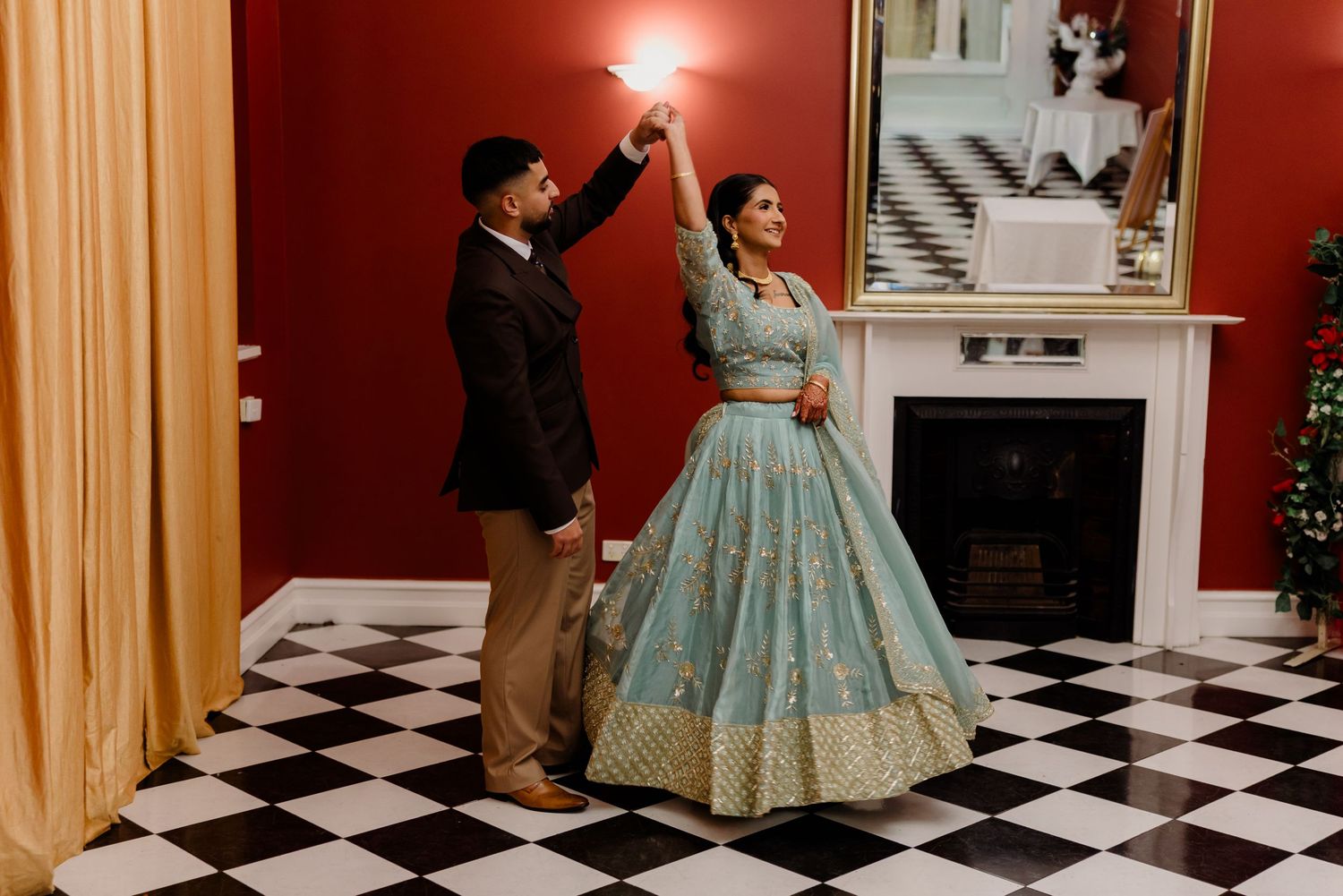 Couple in traditional South Asian attire dancing on checkered floor in elegant red-walled venue.