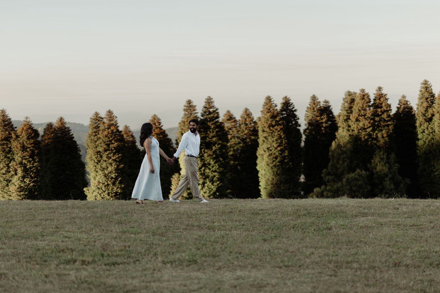 Couple walking hand in hand across a green field with tall evergreen trees in the background at golden hour.