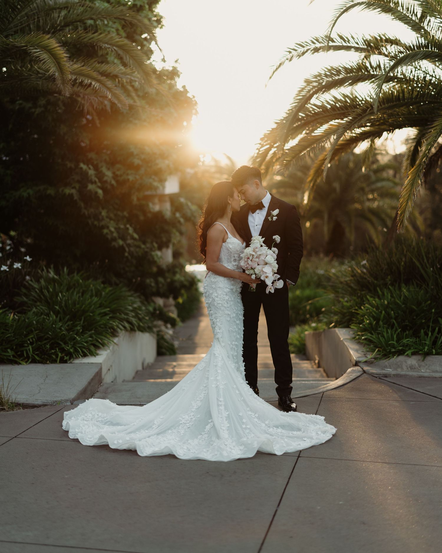 Wedding couple posing at sunset among palm trees in elegant attire.