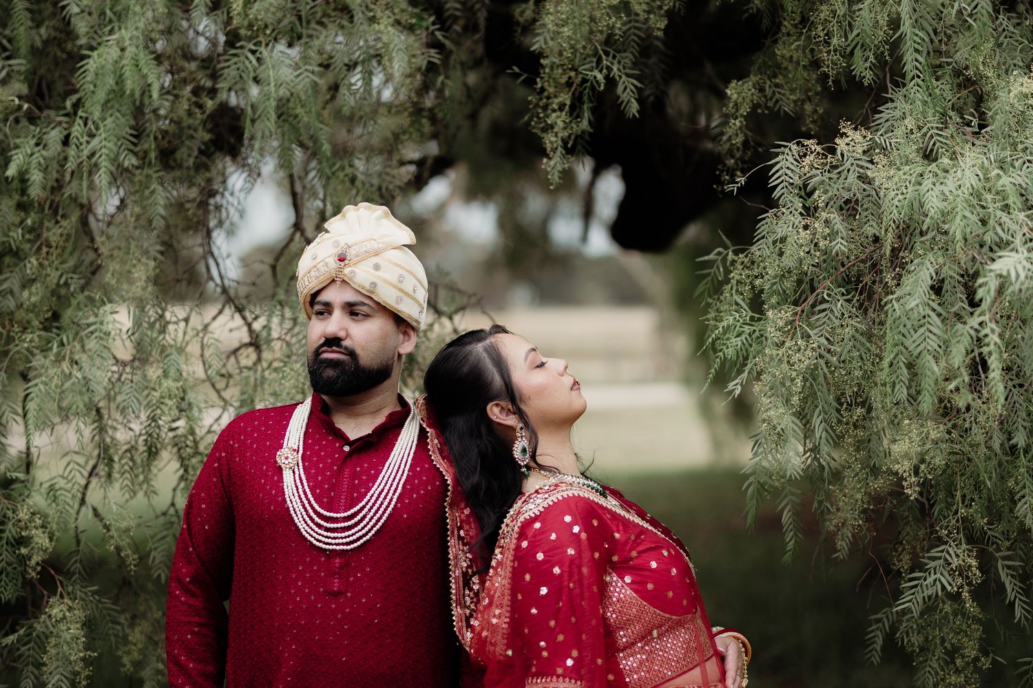 Indian couple in red bridal attire posing under pine trees, groom in turban and bride in saree with pearl jewelry.