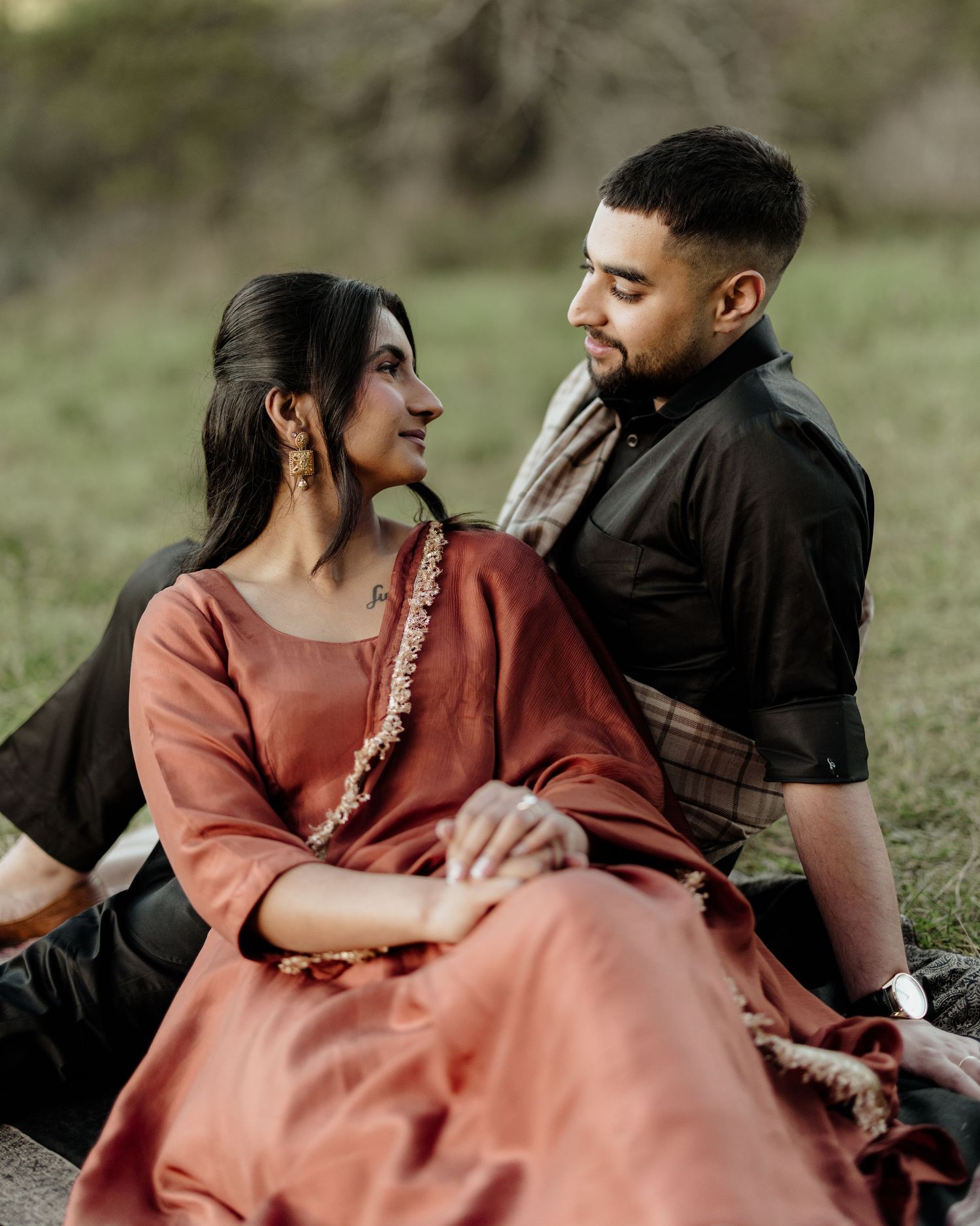 A South Asian couple in traditional attire sits outdoors, sharing an intimate moment, the woman in rust salwar and man in black.