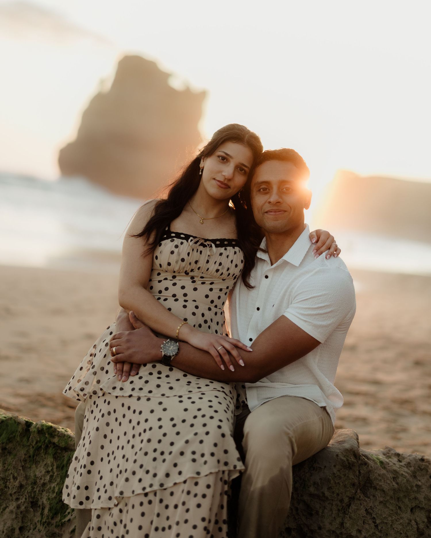 Couple posing romantically on a beach at golden sunset with rocky cliffs in the background.