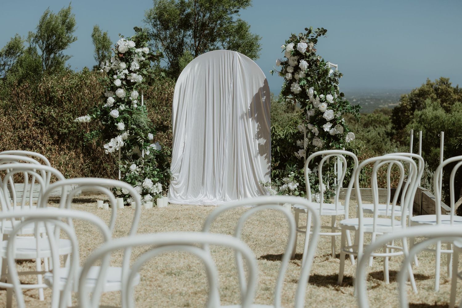 Outdoor wedding ceremony setup with white chairs and floral arrangements under blue sky.