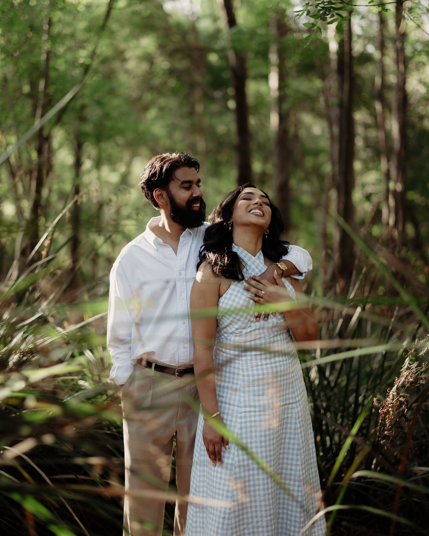 A couple laughing together in a lush green forest, man in white shirt embracing woman in blue gingham dress.