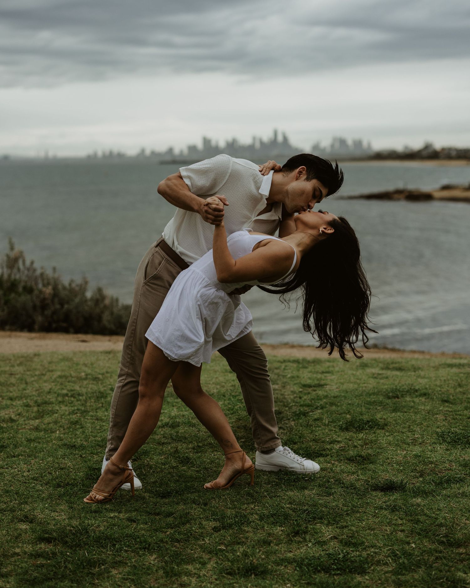 Couple performs romantic dance pose on grass with city skyline visible in background.