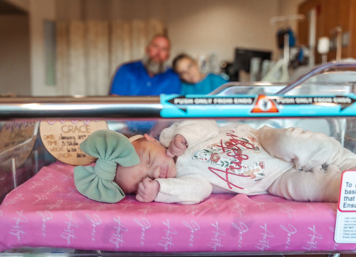 Newborn sleeping peacefully in hospital bassinet with mint green bow parents smiling in background during hospital fresh 48 session