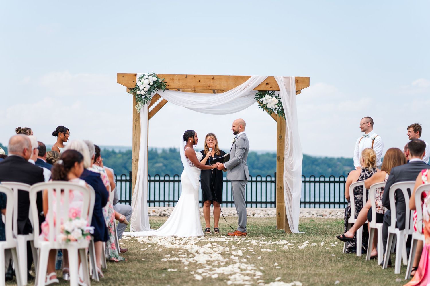 A panoramic view of an outdoor wedding ceremony by the water with white chairs and a wooden arch.
Wedding in Niagara.