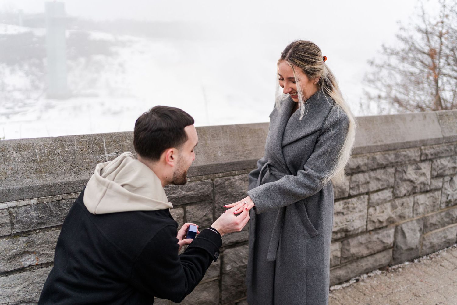 A couple shares a romantic proposal moment on a stone wall during winter with snow in the background.
Proposal captured in Niagara falls.