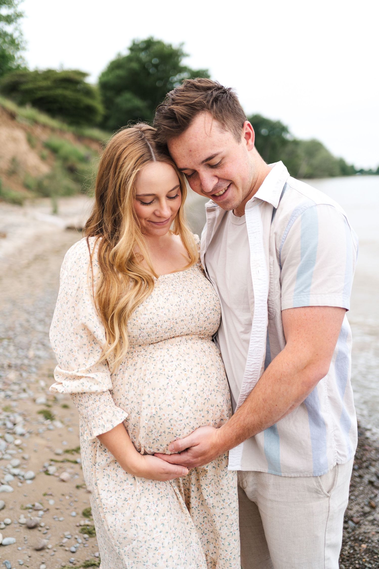 A couple shares an intimate moment on a rocky beach as they celebrate their pregnancy in matching light colored outfits.
Niagara Photos.