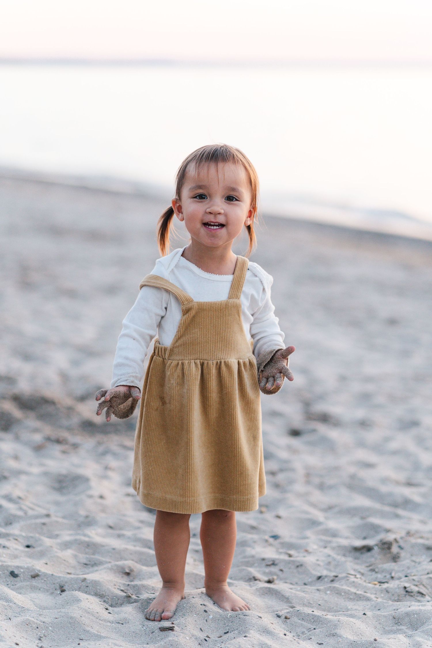 Small child in tan overalls plays barefoot on sandy beach at sunset.
Niagara family photos.