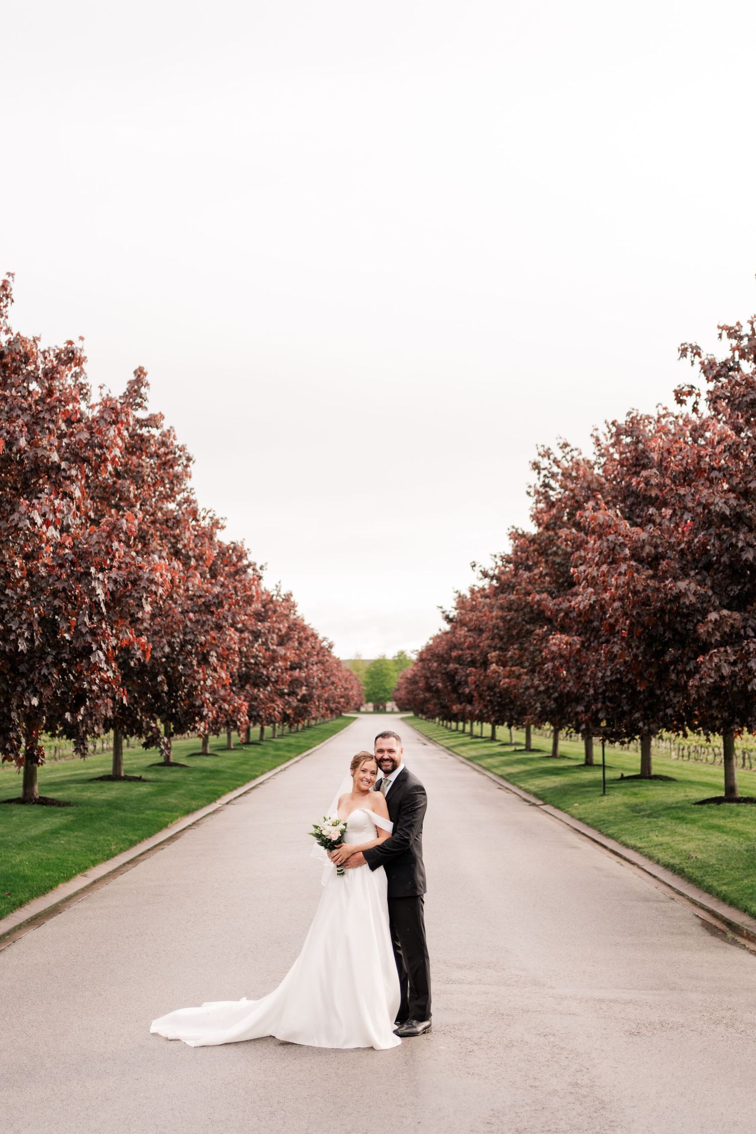 Wedding couple embracing on tree-lined road with pink blossoms.
Niagara wedding photos.