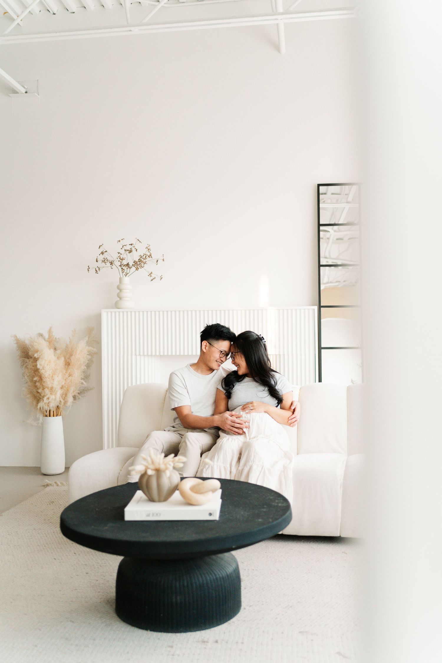 A couple sits together on a white couch in a minimalist room with pampas grass decor and a round black coffee table.