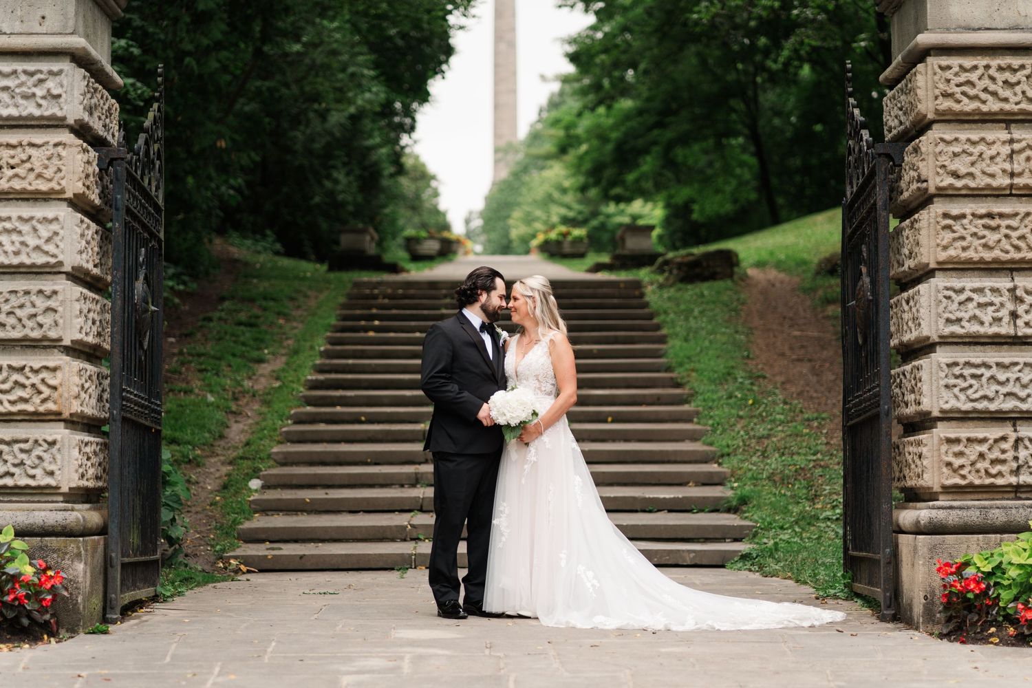 Bride and groom share a romantic moment under stone archway with grand staircase and monument backdrop.