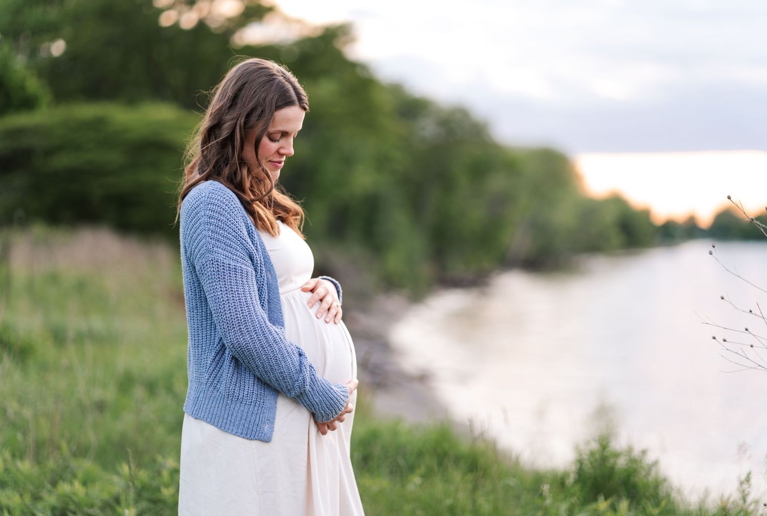 An expectant mother in a blue cardigan gazes down at her baby bump by a serene waterfront at sunset.
Niagara maternity photos.