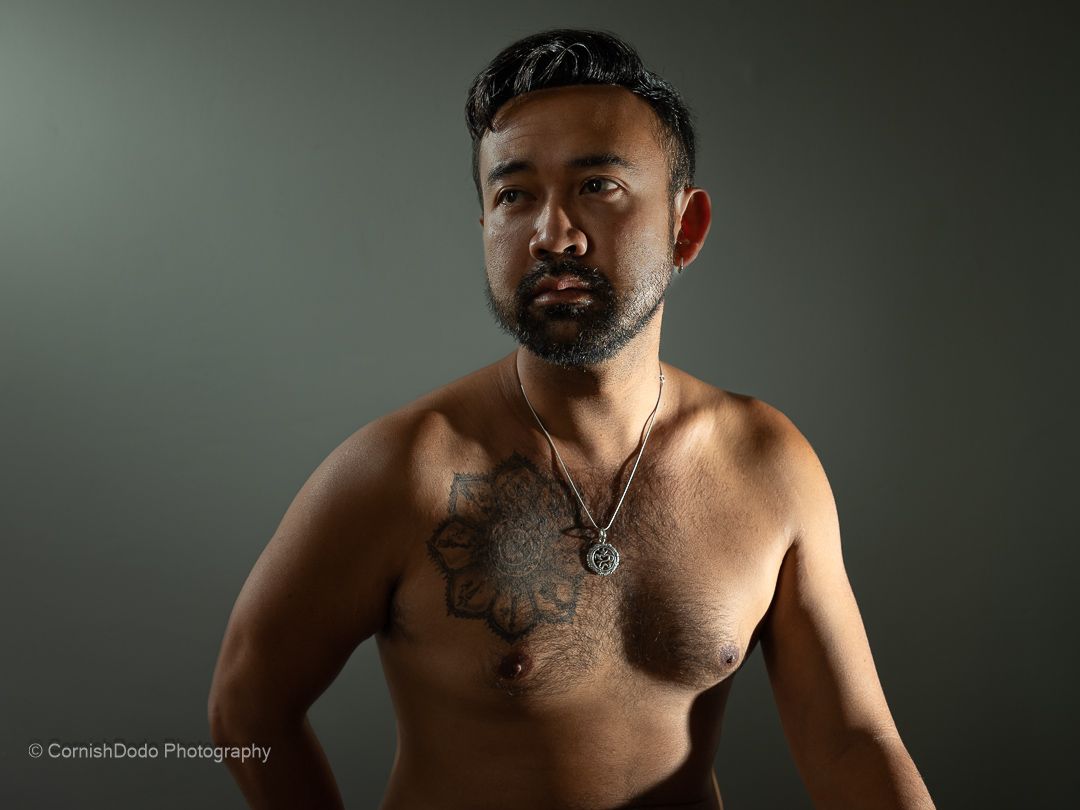 Asian man, shirtless, sitting in studio light, part of a portrait series exploring masculinity and body‑positive representation.