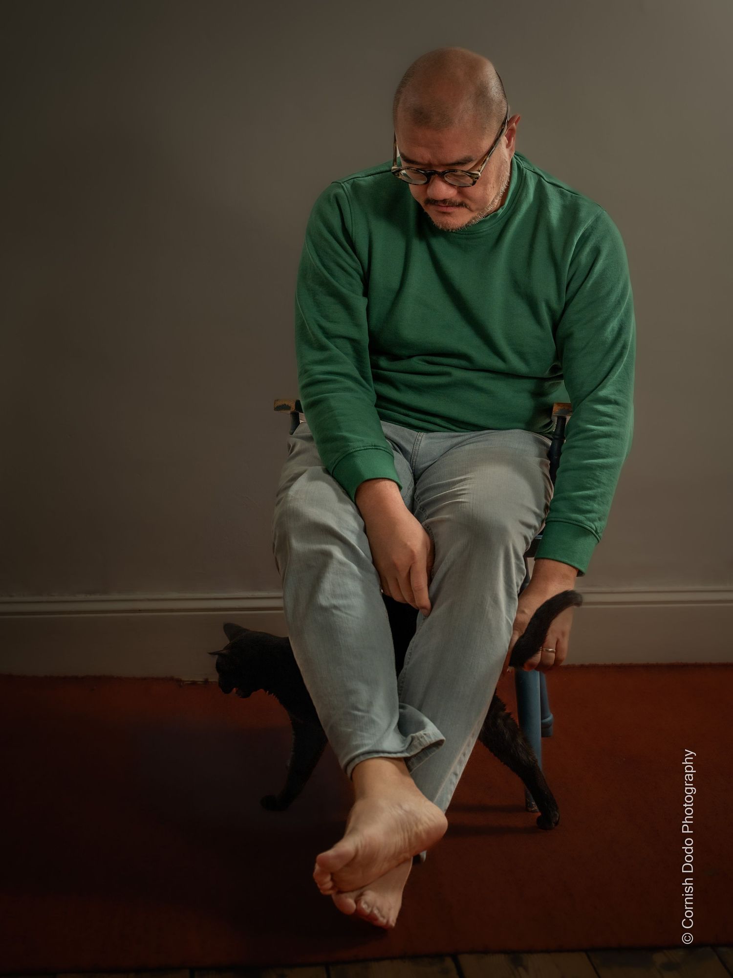 Asian man, clothed, sitting in studio light, part of a portrait series exploring masculinity and body‑positive representation.