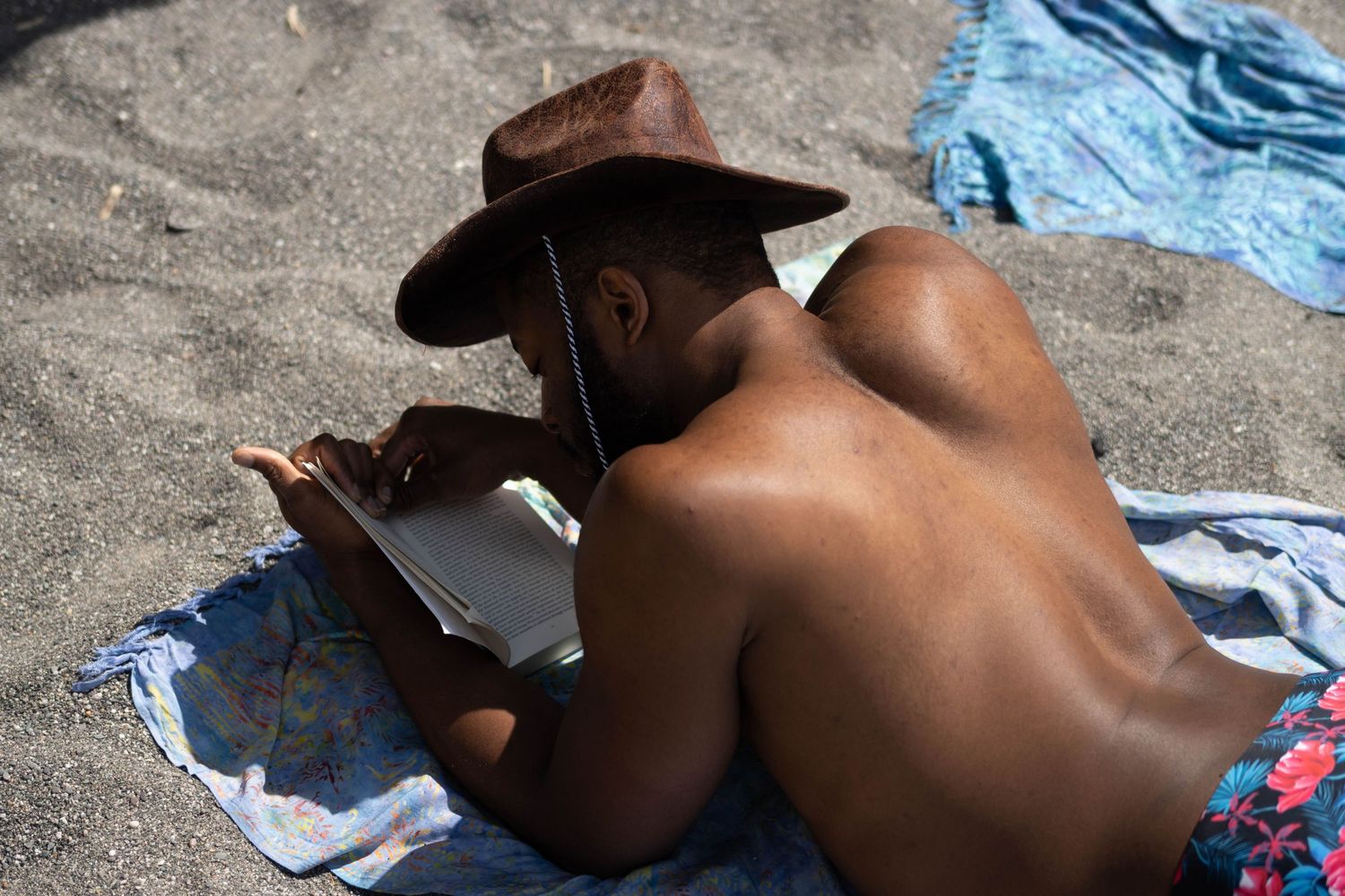 A Black man is lying shirtless on a beach, wearing a hat and reading a book. part of a masculinity/identity portrait series.