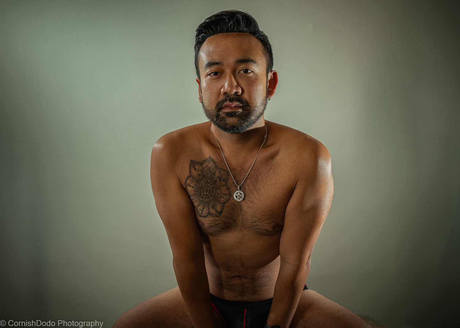 Asian man, shirtless, sitting in studio light, part of a portrait series exploring masculinity and body‑positive representation.
