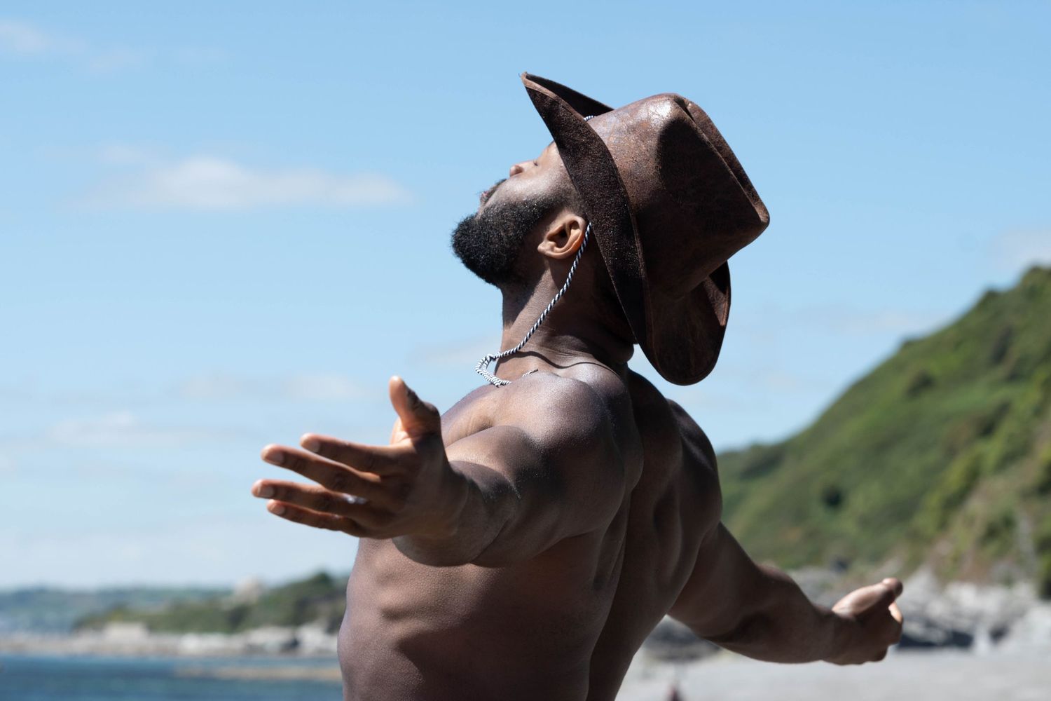 Black man in a cowboy hat standing shirtless with arms outstretched in a sunny outdoor landscape, part of a masculinity portrait series.