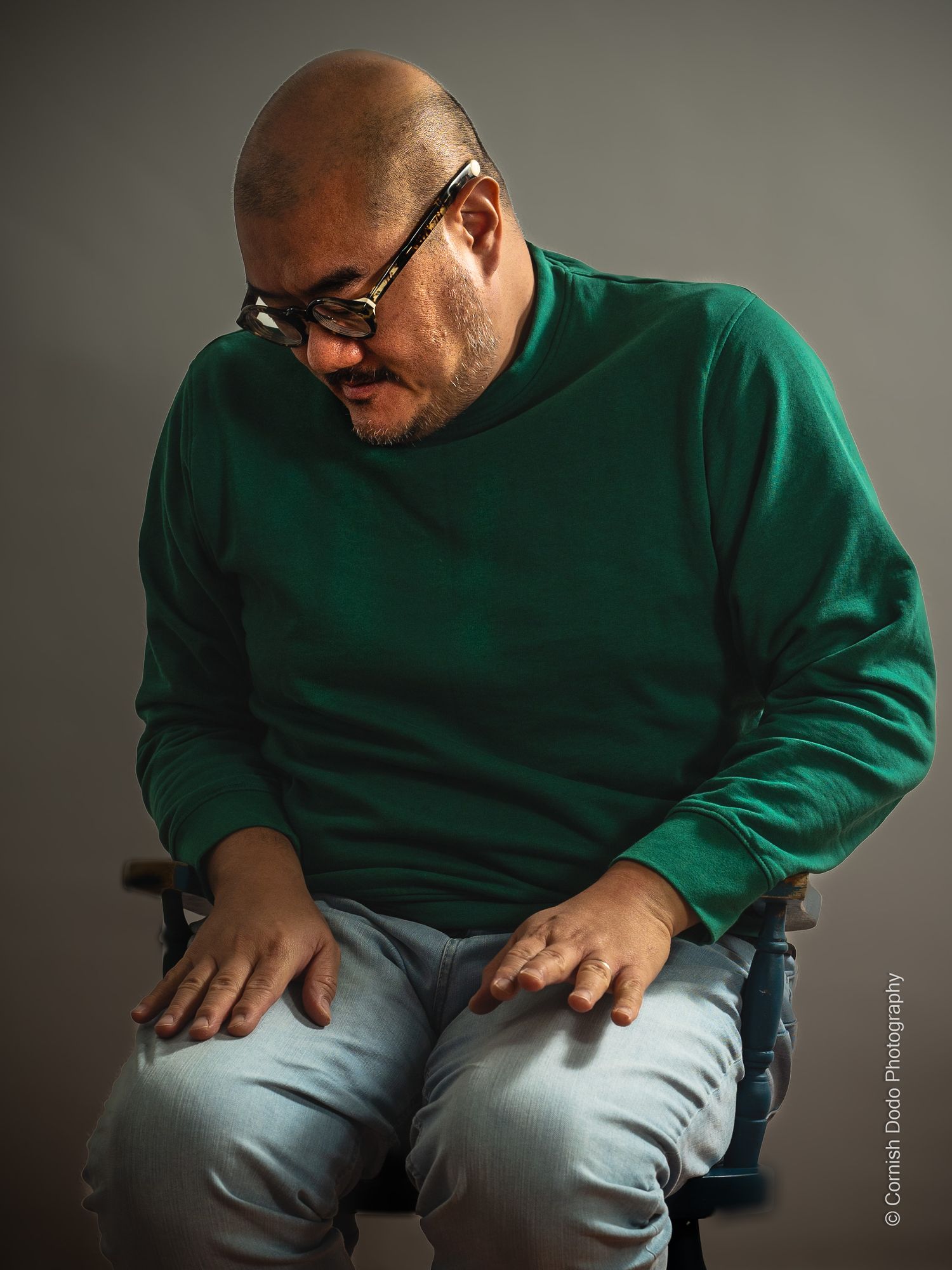 Asian man, clothed, sitting in studio light, part of a portrait series exploring masculinity and body‑positive representation.