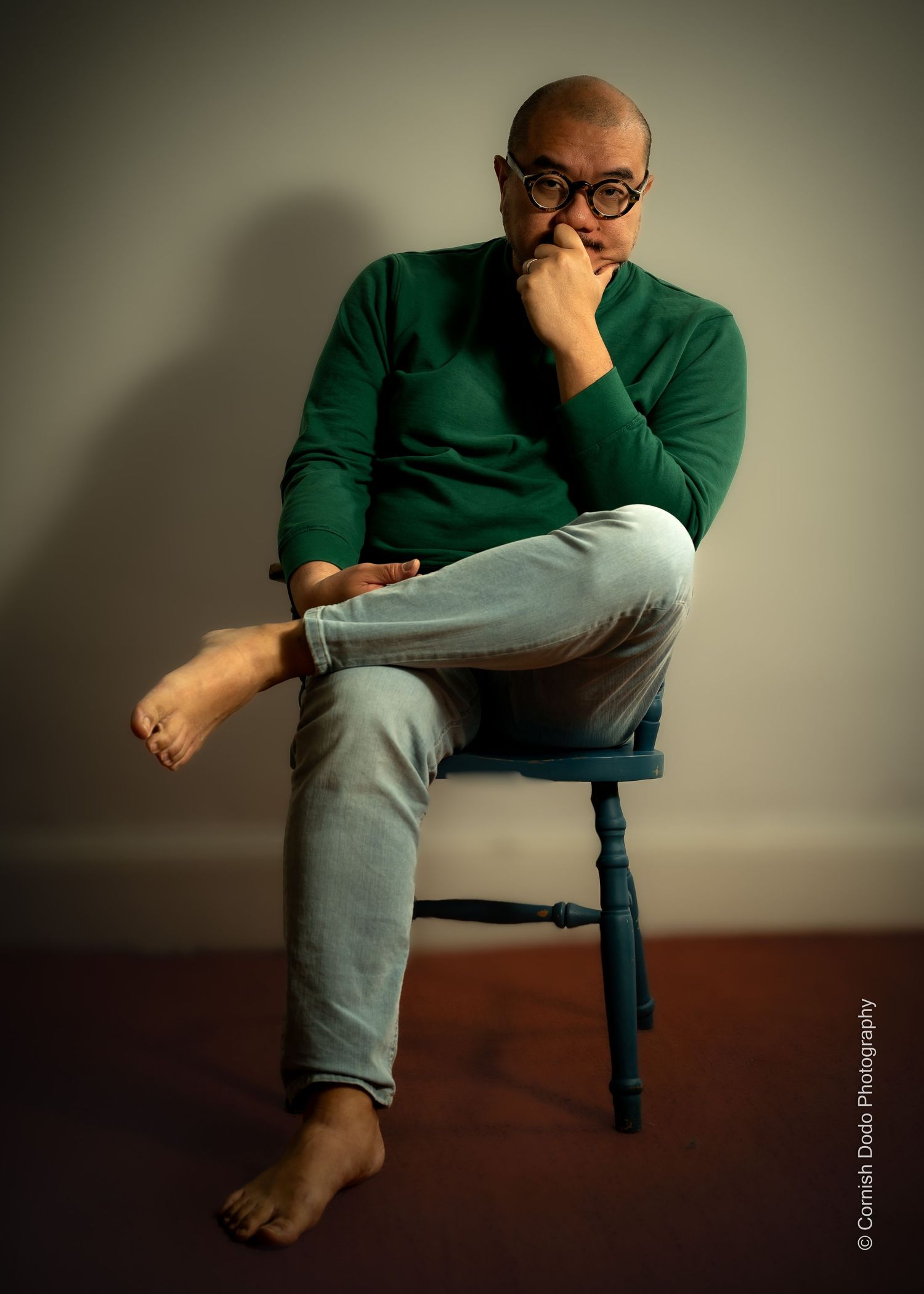 Asian man, clothed, sitting in studio light, part of a portrait series exploring masculinity and body‑positive representation.