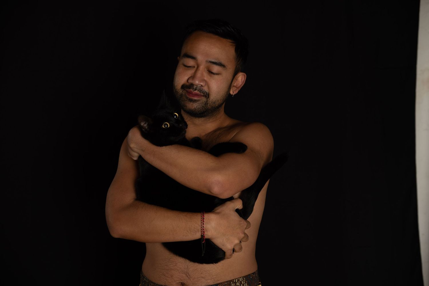 Asian man, shirtless, standing with cat in studio light, part of a portrait series exploring masculinity and body‑positive representation.