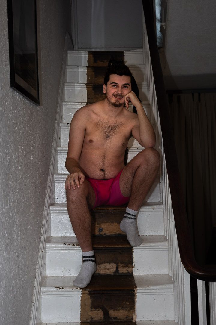 White man, wearing only shorts, seated in natural light, part of a portrait series exploring masculinity and body‑positive representation.
