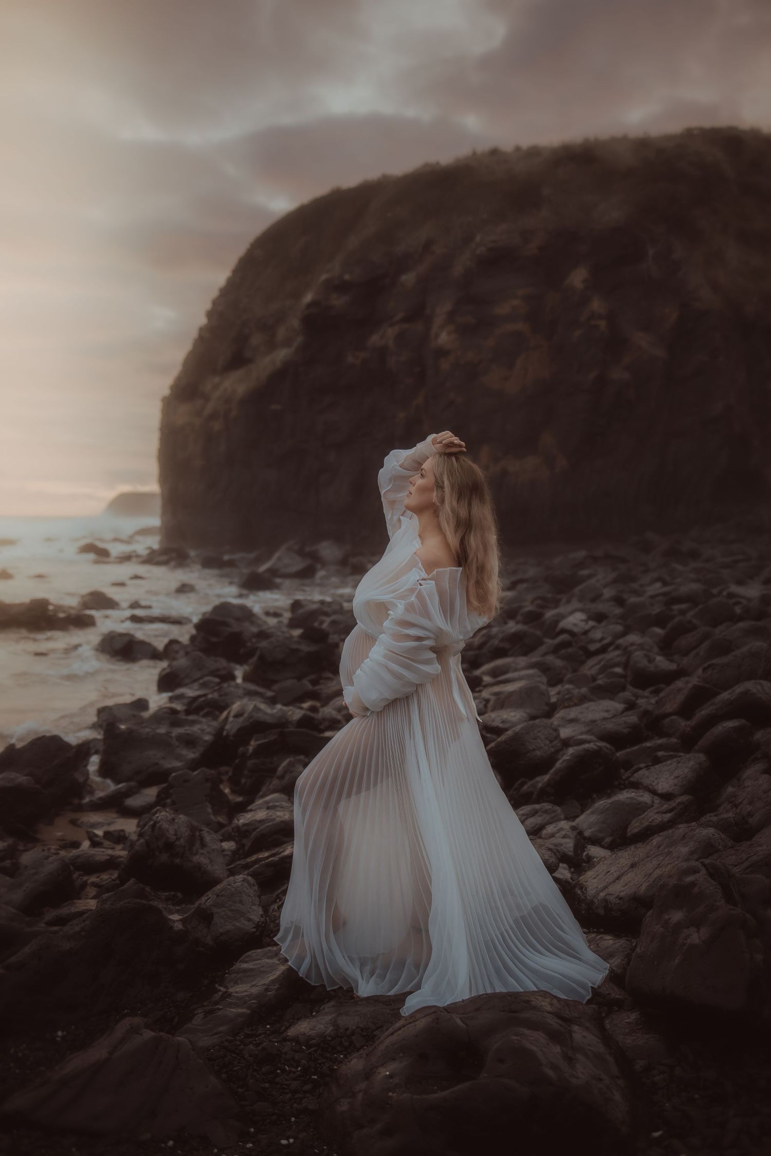 A dramatic coastal scene with a flowing white dress against rocky shores at sunset.
