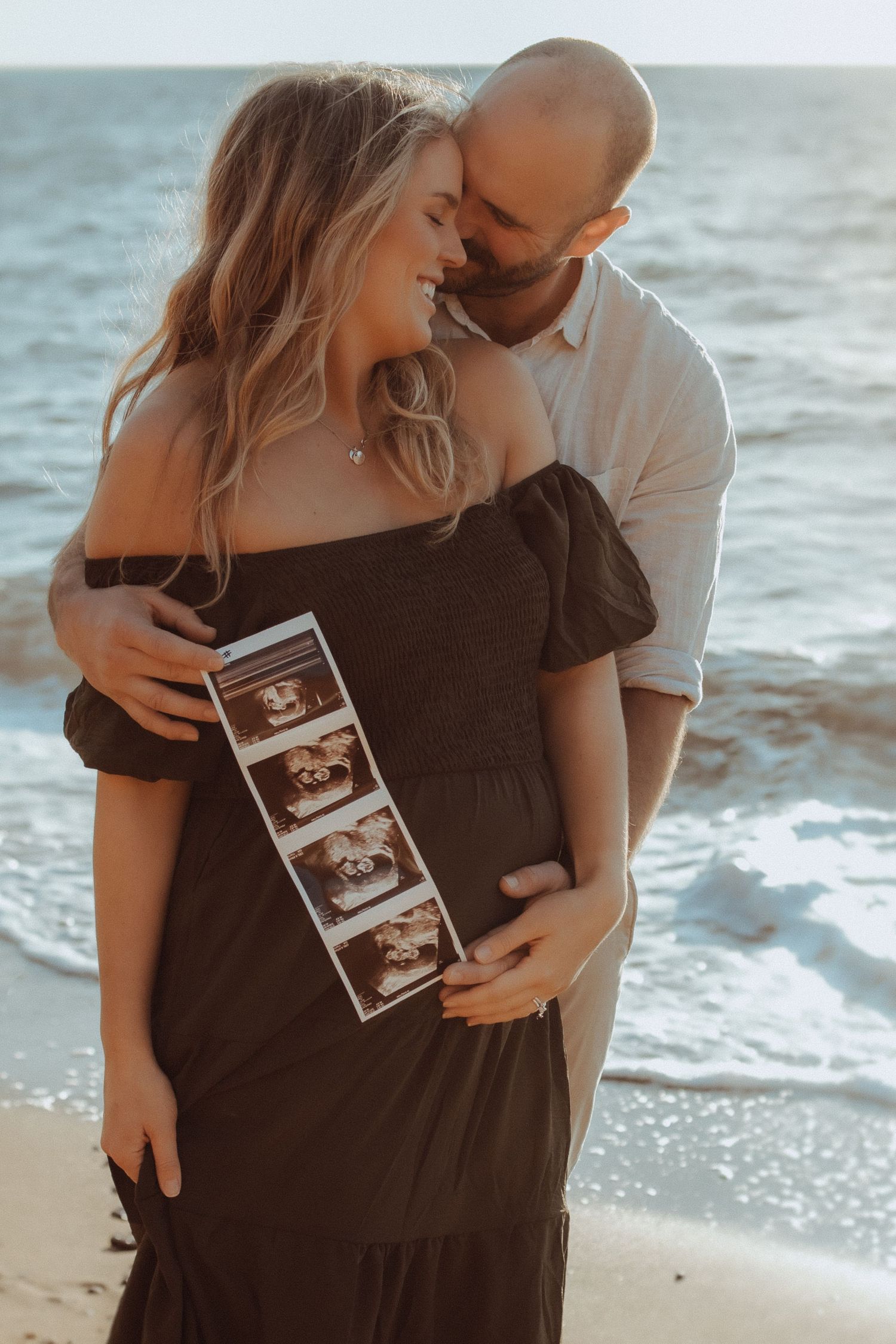 Couple holding ultrasound photos on beach during sunset pregnancy announcement shoot.