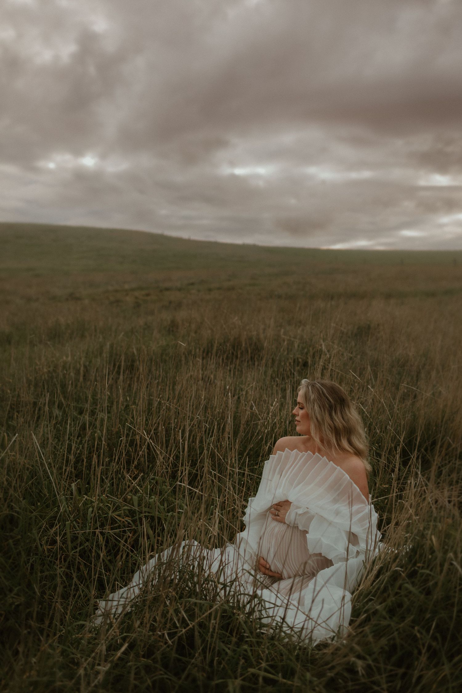 A maternity portrait in tall grass with moody clouds overhead.