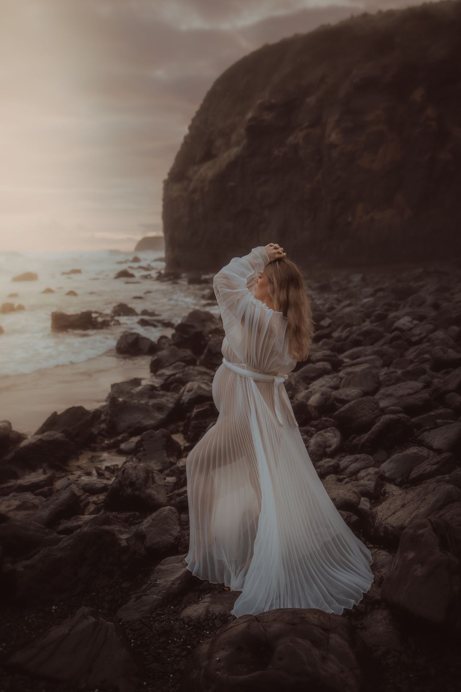 An ethereal white gown catches the wind against rocky coastal cliffs at dusk.