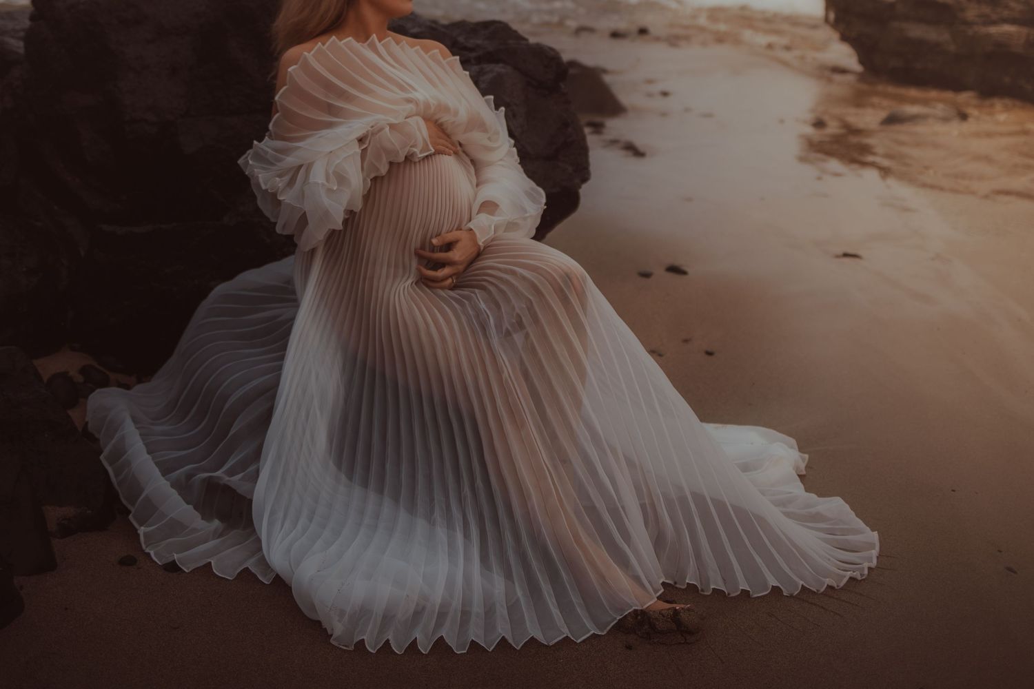 A pleated white dress flows elegantly across dark rocks at sunset on a beach.
