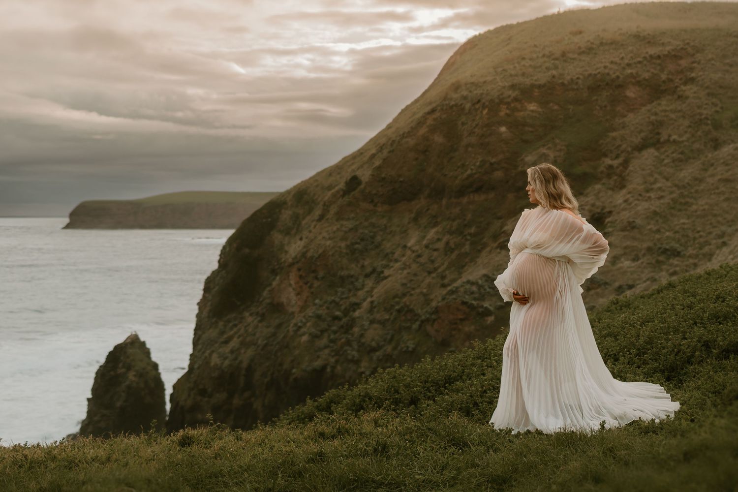 A flowing white dress billows in the wind against dramatic coastal cliffs and ocean views at sunset.