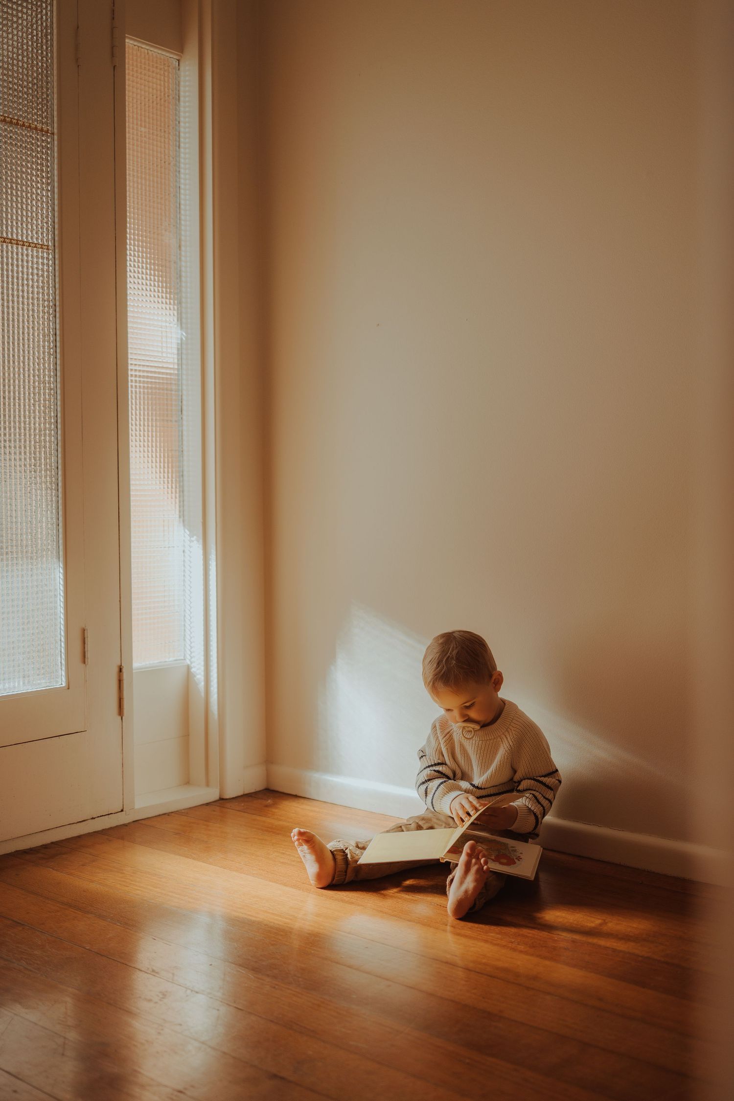 Child reading book alone in sunlit corner on hardwood floor.