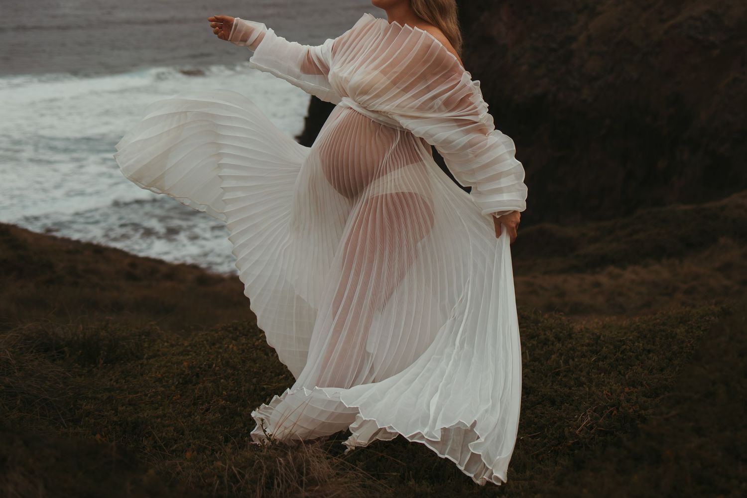 Ethereal white pleated dress flowing in motion against dramatic coastal backdrop.