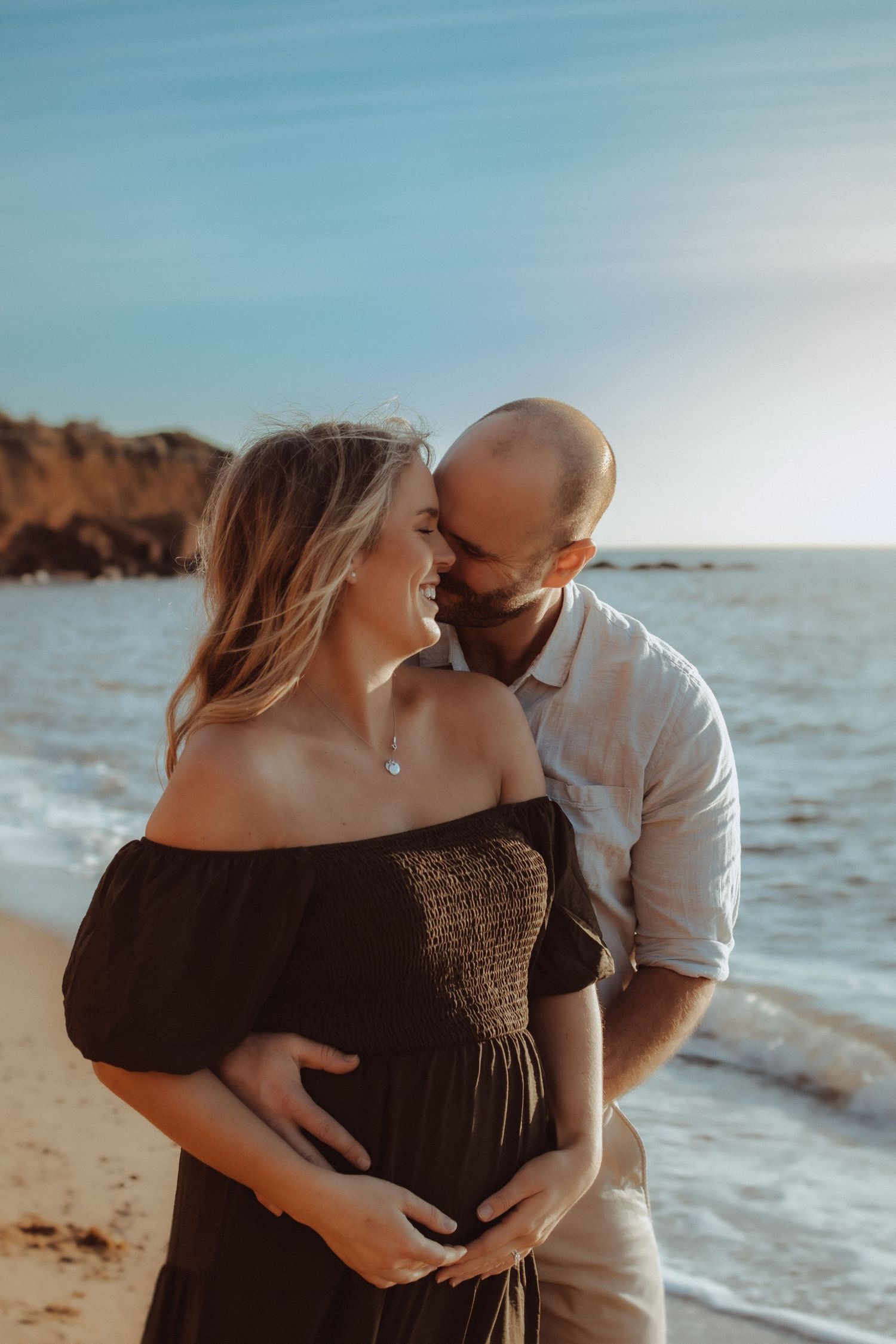 Couple embracing on a sandy beach at sunset with waves crashing in the background.